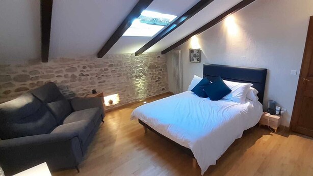 Une chambre avec un mur en pierre, un lit avec une literie blanche, un fauteuil gris et une porte en bois.