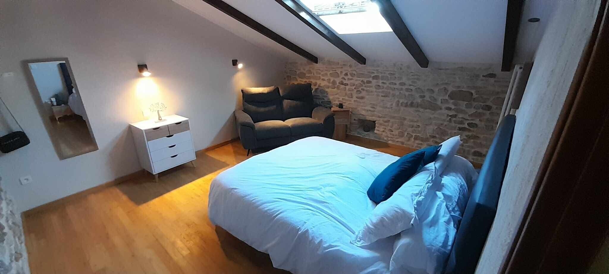 A bedroom with a bed, a chair, a nightstand, a mirror, and a stone wall.