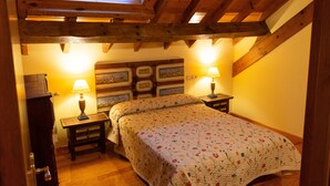 Un dormitori amb sostre de fusta, un llit amb roba de llit floral, dues làmpades de nit i una taula petita amb una làmpada.