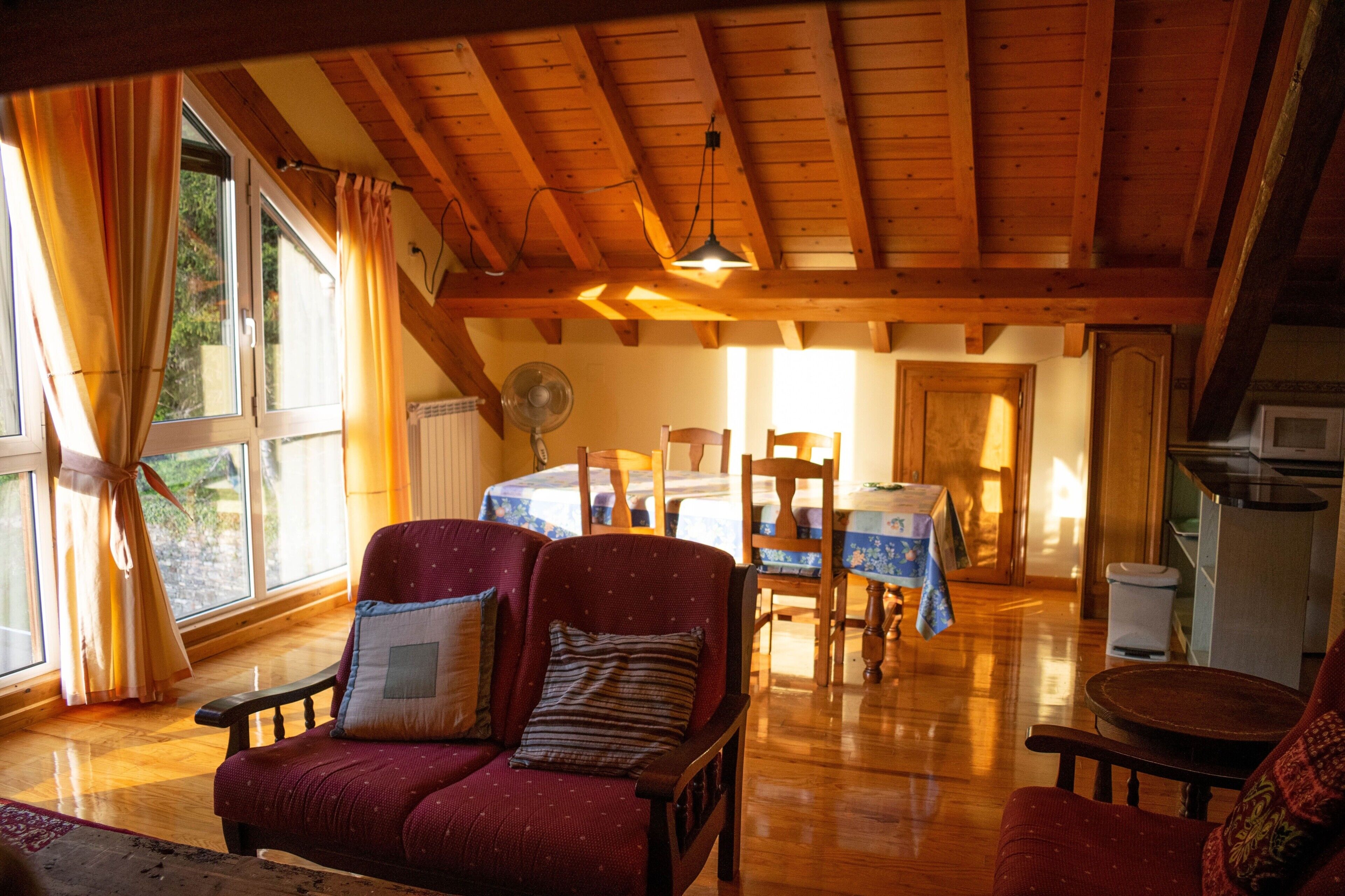 A cozy room with a wooden ceiling, a dining table with chairs, a red armchair, and a small table with a lamp.