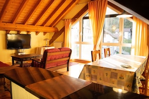 Una habitación acogedora con techo de madera, televisor, una mesa de comedor con mantel floral y una ventana con cortinas.