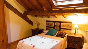 Un dormitori amb sostre de fusta, una clau sense tallar, un llit amb roba de llit floral, una tauleta de nit amb una làmpada i una paret amb quadres emmarcats.