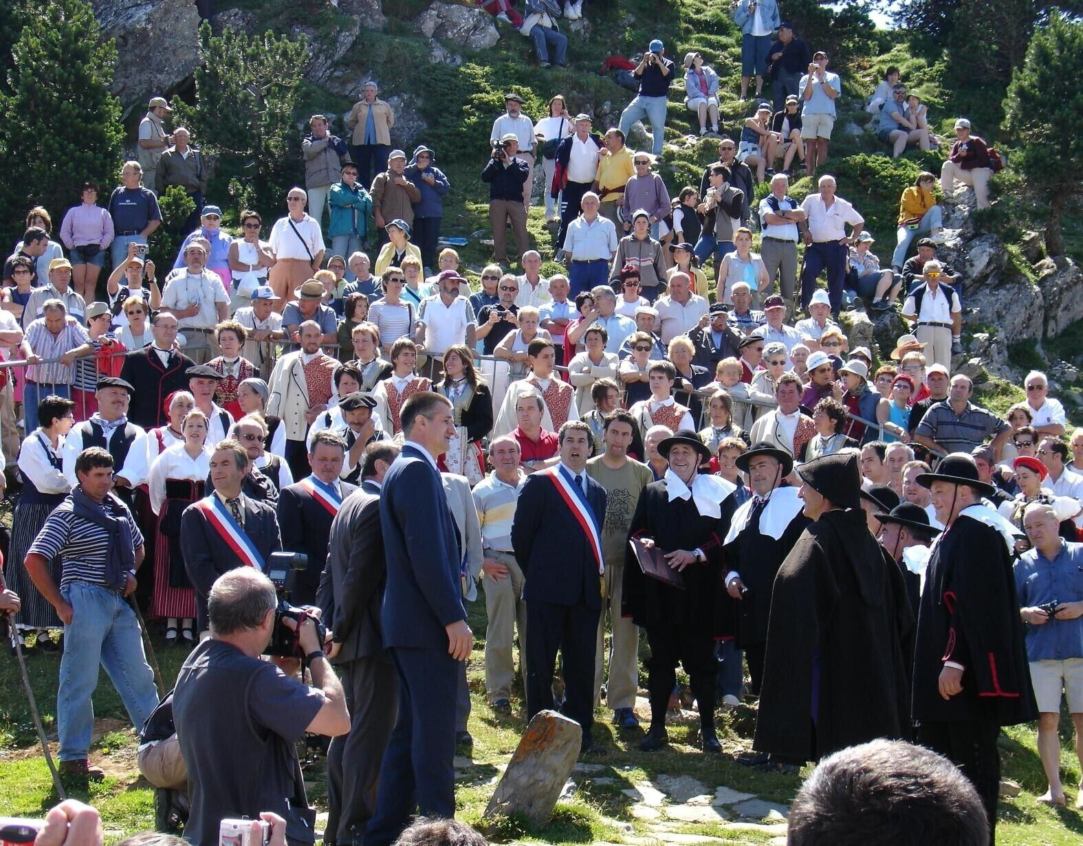 A large outdoor gathering of people, some in traditional attire, on a grassy slope with trees and rocky terrain in the background.