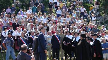 A large outdoor gathering of people, some in traditional attire, on a grassy slope with trees and rocky terrain in the background.
