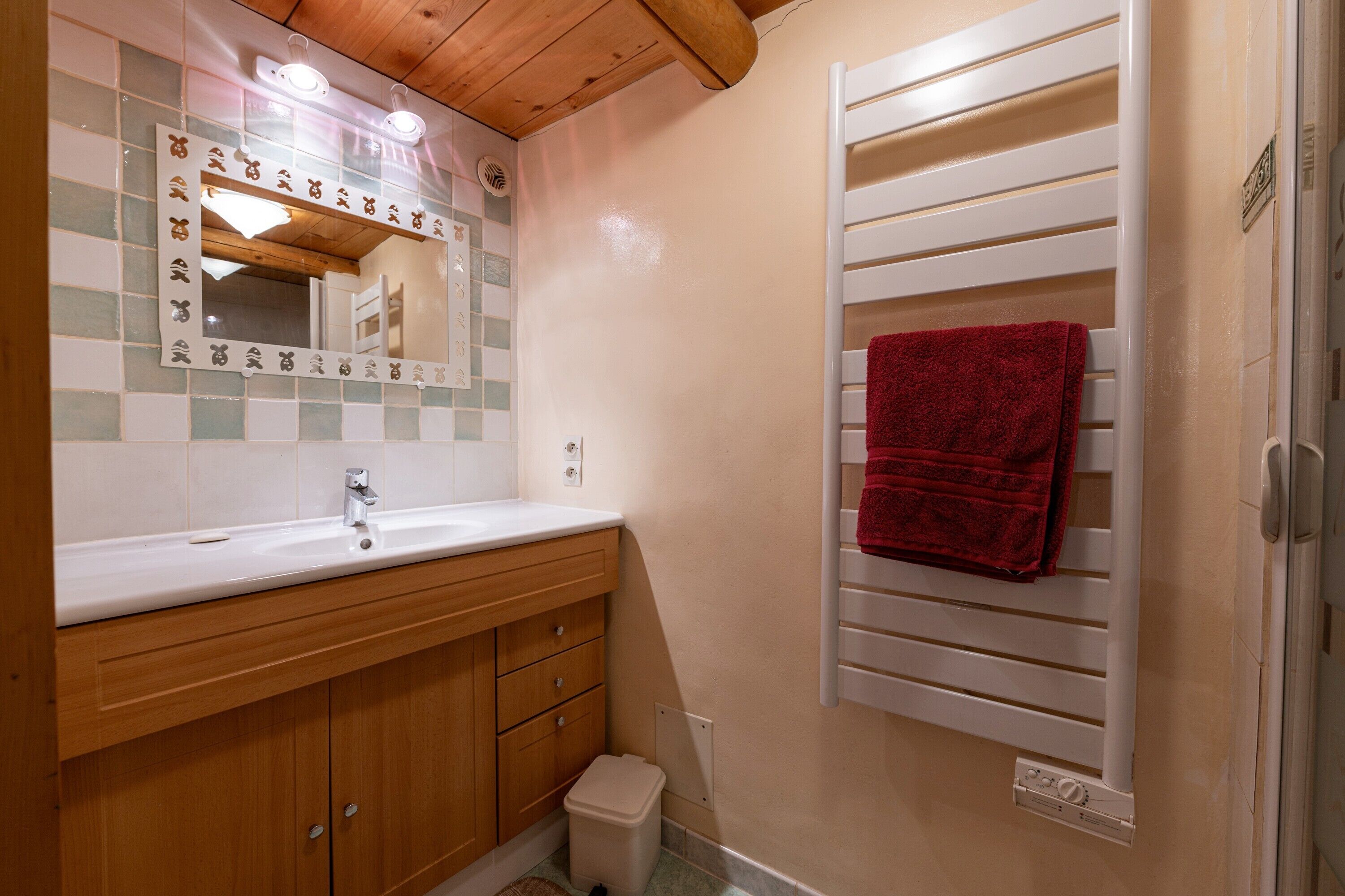 Une salle de bain avec un meuble-lavabo en bois, un miroir orné de carreaux décoratifs, un porte-serviettes avec une serviette rouge et un radiateur blanc.