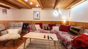 Ein gemütliches Zimmer mit Holzbalkendecke, einem Bett mit gemusterten Betttextilien, einem Sessel mit Felldecke und einem Tisch mit Fernbedienungen.