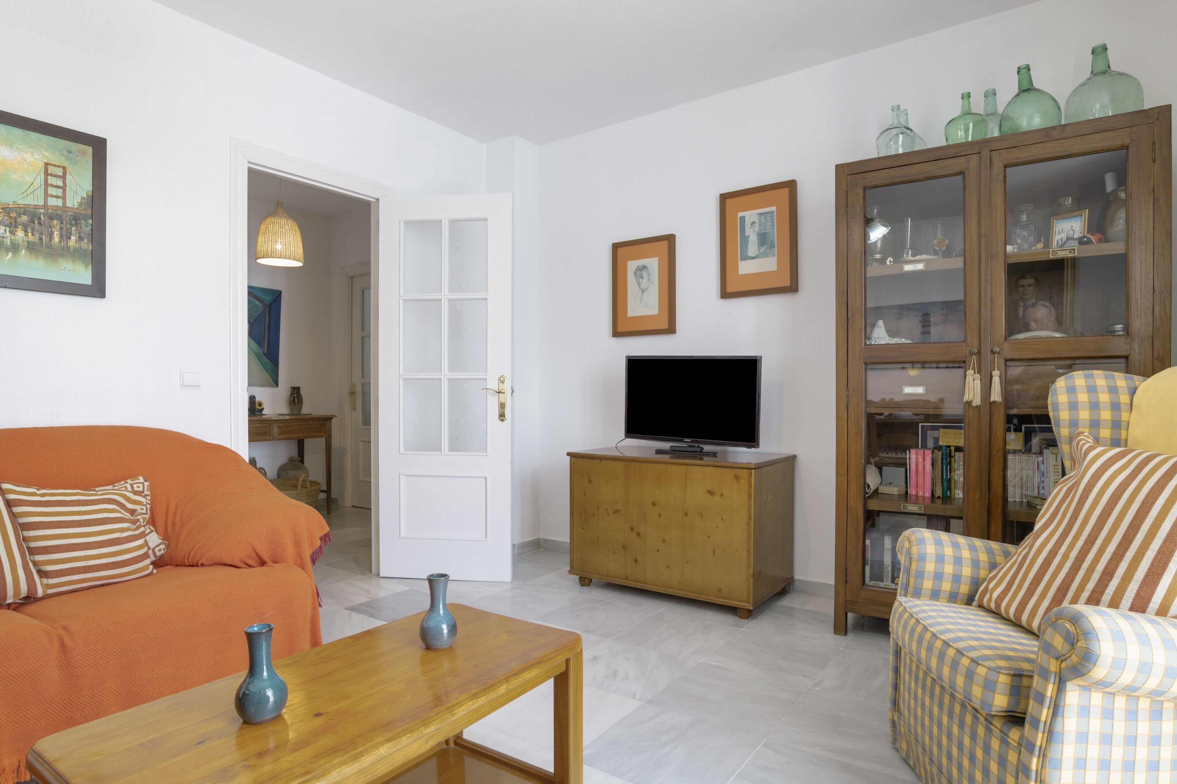Una sala de estar con un sofá, un televisor sobre un mueble de madera, una mesa con un florero y un sillón a cuadros.