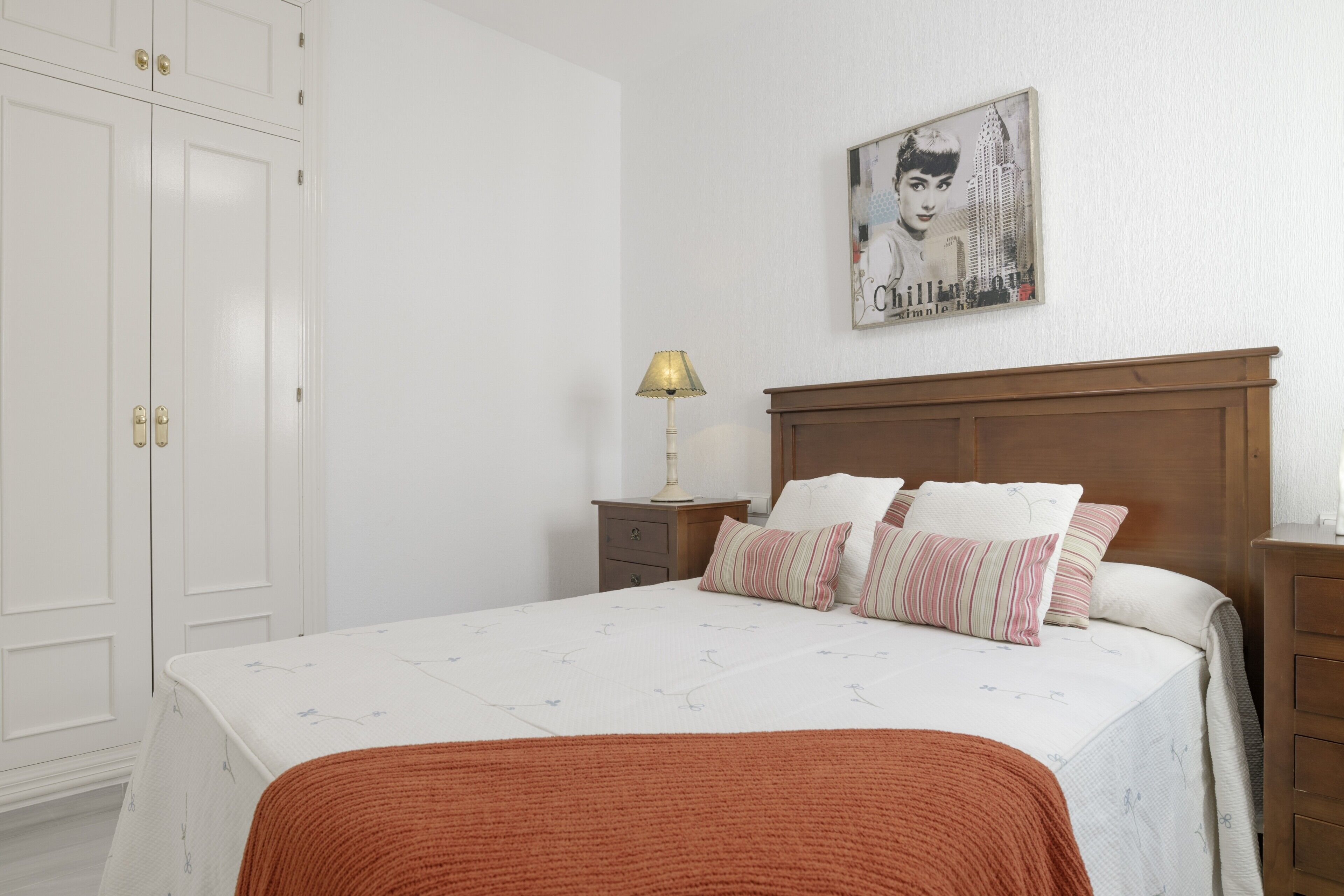 A bedroom with a bed, a nightstand, a lamp, a wardrobe, and a wall-mounted picture.