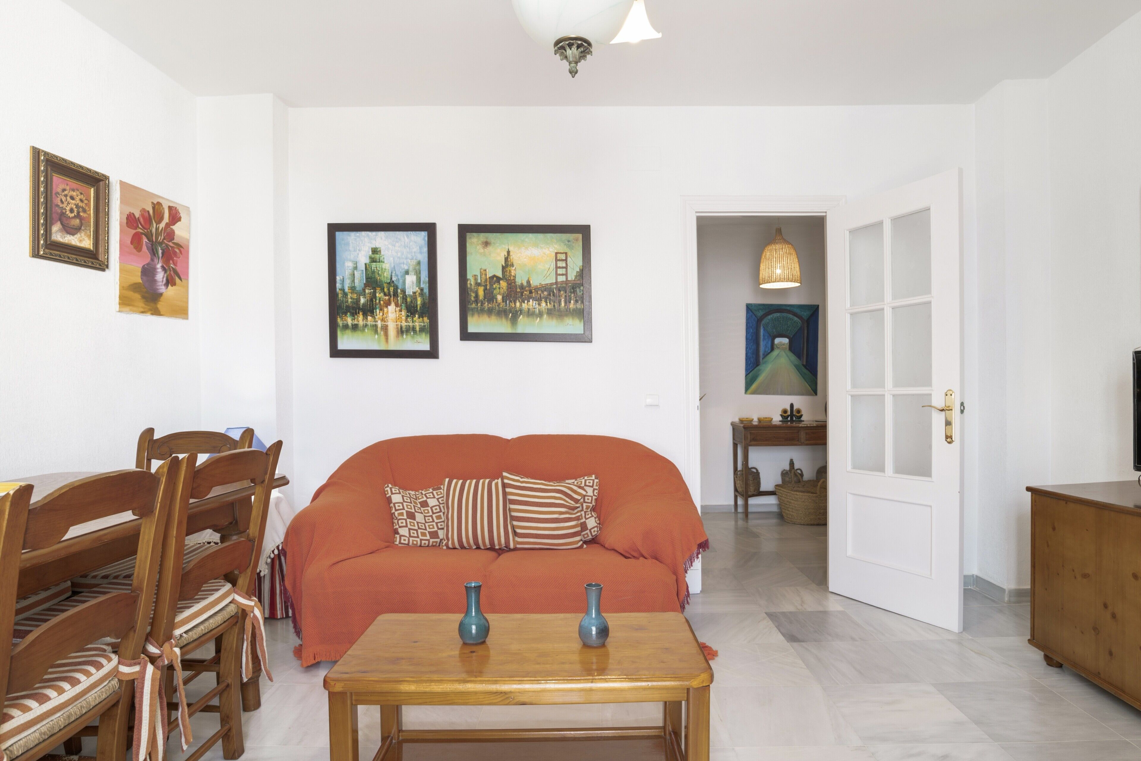 A living room with a sofa, wooden chairs, a table with vases, and framed pictures on the wall.