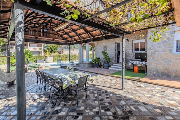 An outdoor patio with a covered seating area, stone flooring, and a view of a stone building in the background.