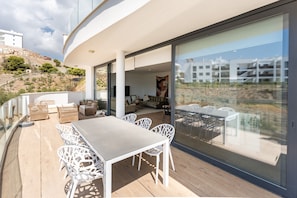 A modern outdoor patio with a dining table and chairs, a sofa, and a large glass wall offering a view of the surrounding landscape. - Stunning Sea View Apartment in Higueron West (Málaga)
