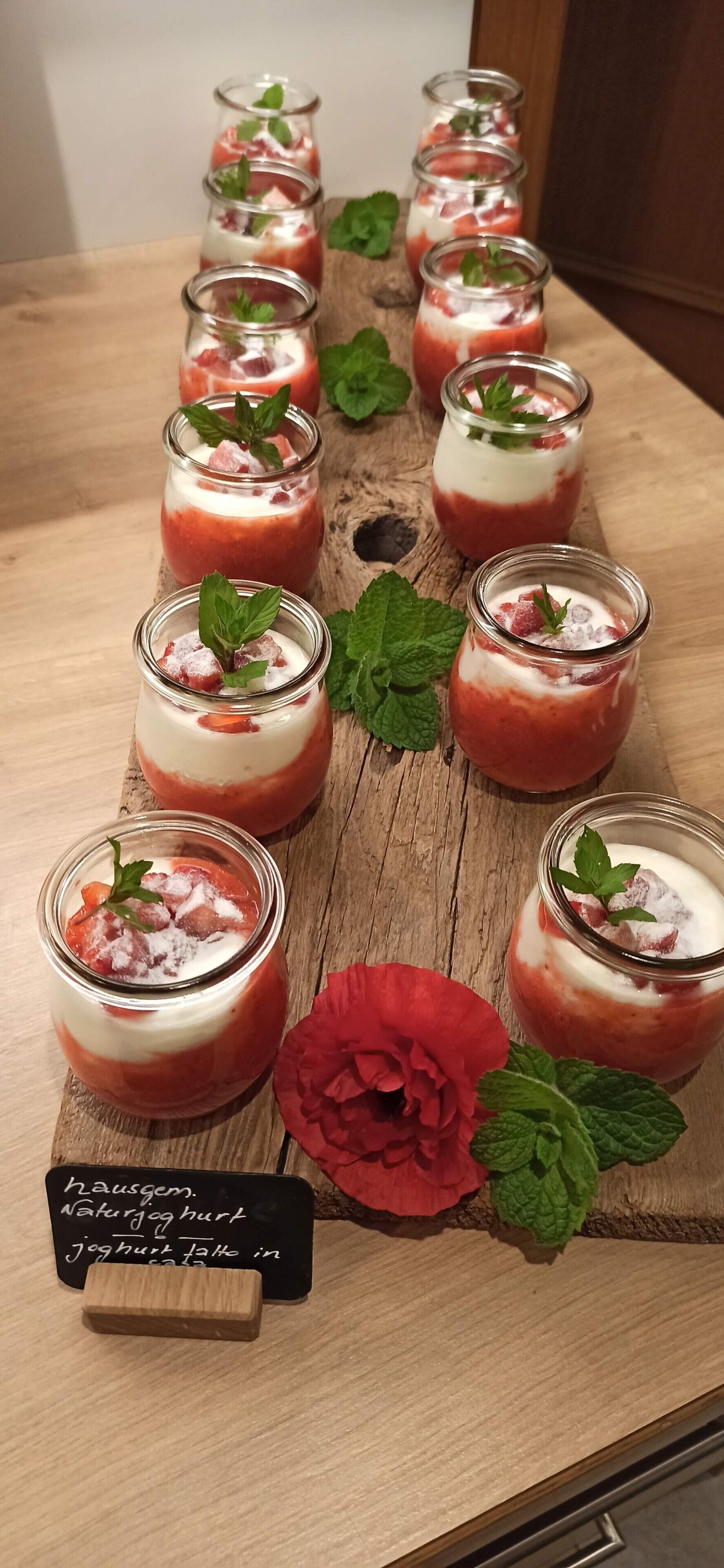 Een houten plank met meerdere glazen potten gevuld met gelaagde desserts, versierd met muntblaadjes en een rode bloem.