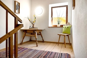 Una escalera de madera, una mesita de madera con un jarrón de flores, una ventana con vistas y un cojín verde sobre un taburete de madera.