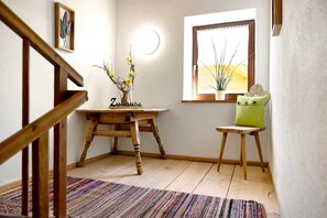Una escalera de madera, una mesita de madera con un jarrón de flores, una ventana con vistas y un cojín verde sobre un taburete de madera.