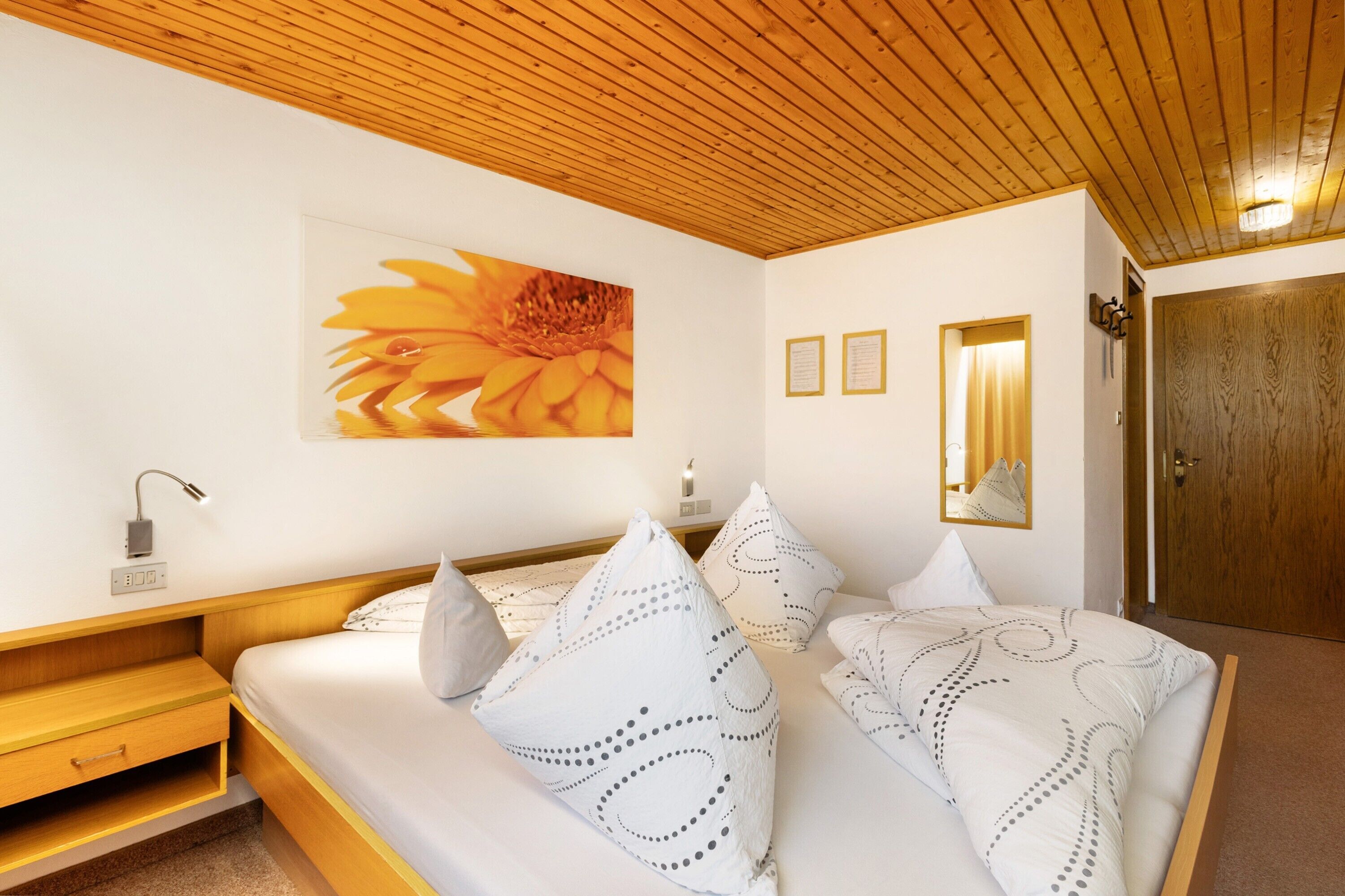 Une chambre avec un plafond en bois, un lit agrémenté d’oreillers à motifs, une lampe fixée au mur et un tableau encadré.