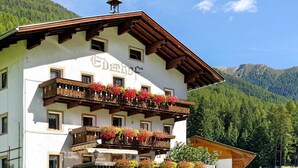 Un edifici de diverses plantes amb balcons, una torre campanar i un paisatge de muntanyes al fons.