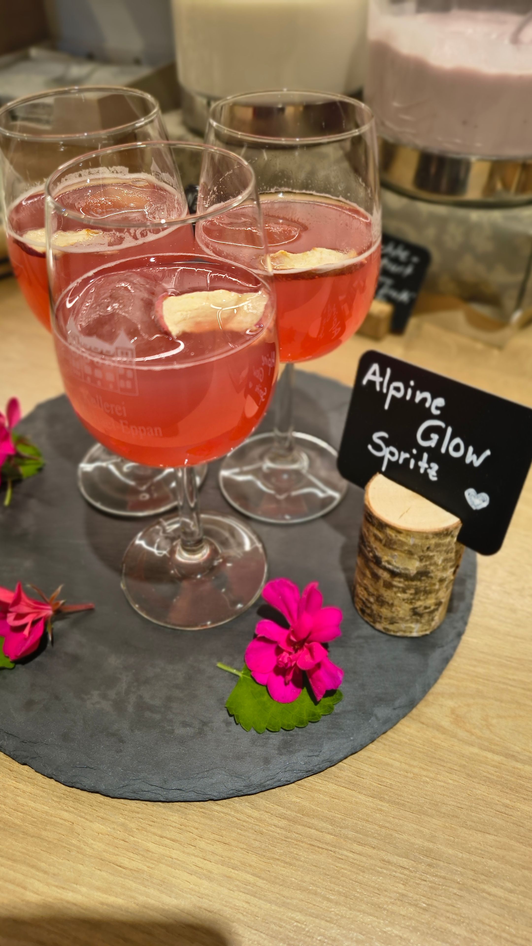 Un tavolo con quattro bicchieri di una bevanda rossa, un cartello con la scritta 'Alpine Glow Spritz' e fiori decorativi.