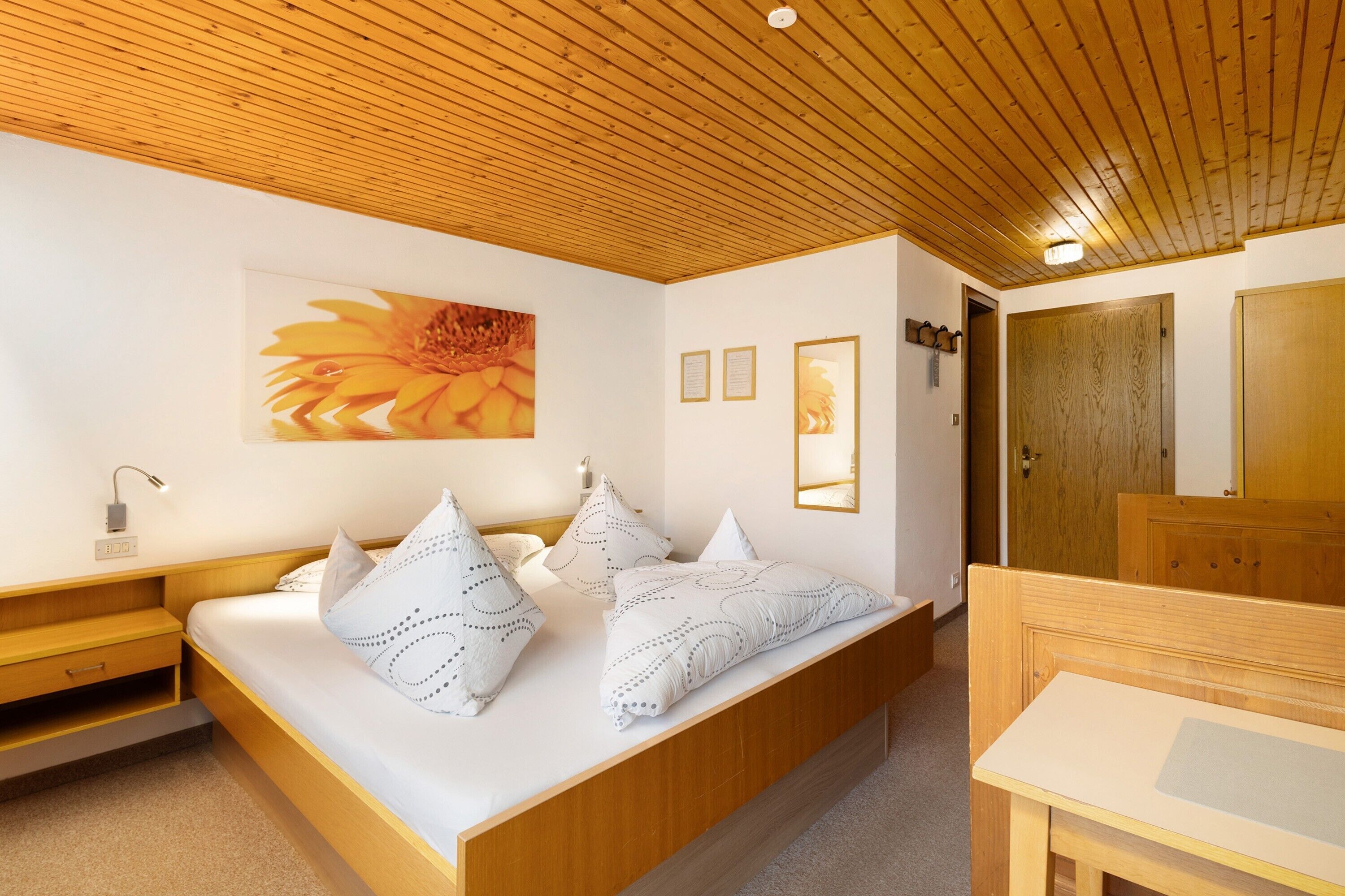 Une chambre avec un plafond en bois, un lit agrémenté d’oreillers à motifs, une table de chevet et une commode, ainsi qu’un miroir.