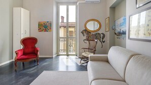 Ein Zimmer mit einem roten Sessel, einem weißen Sofa, einem Schaukelstuhl, einer Chandelüster und einem großen Fenster mit Blick auf die Stadt.