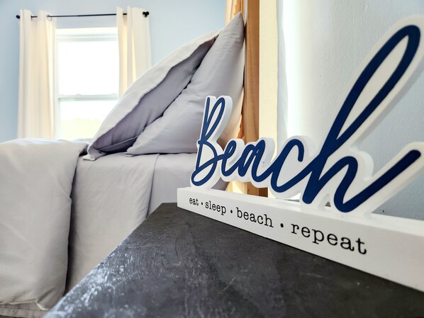 A bed with white bedding and a decorative sign that reads "Beach" with the words "eat sleep beach repeat" underneath.
