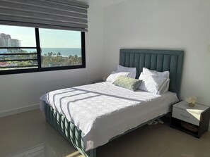 A bedroom with a bed, a nightstand, a window with a view of the ocean and buildings, and a green headboard. - Condo in El Murcielago Beach (Manta)