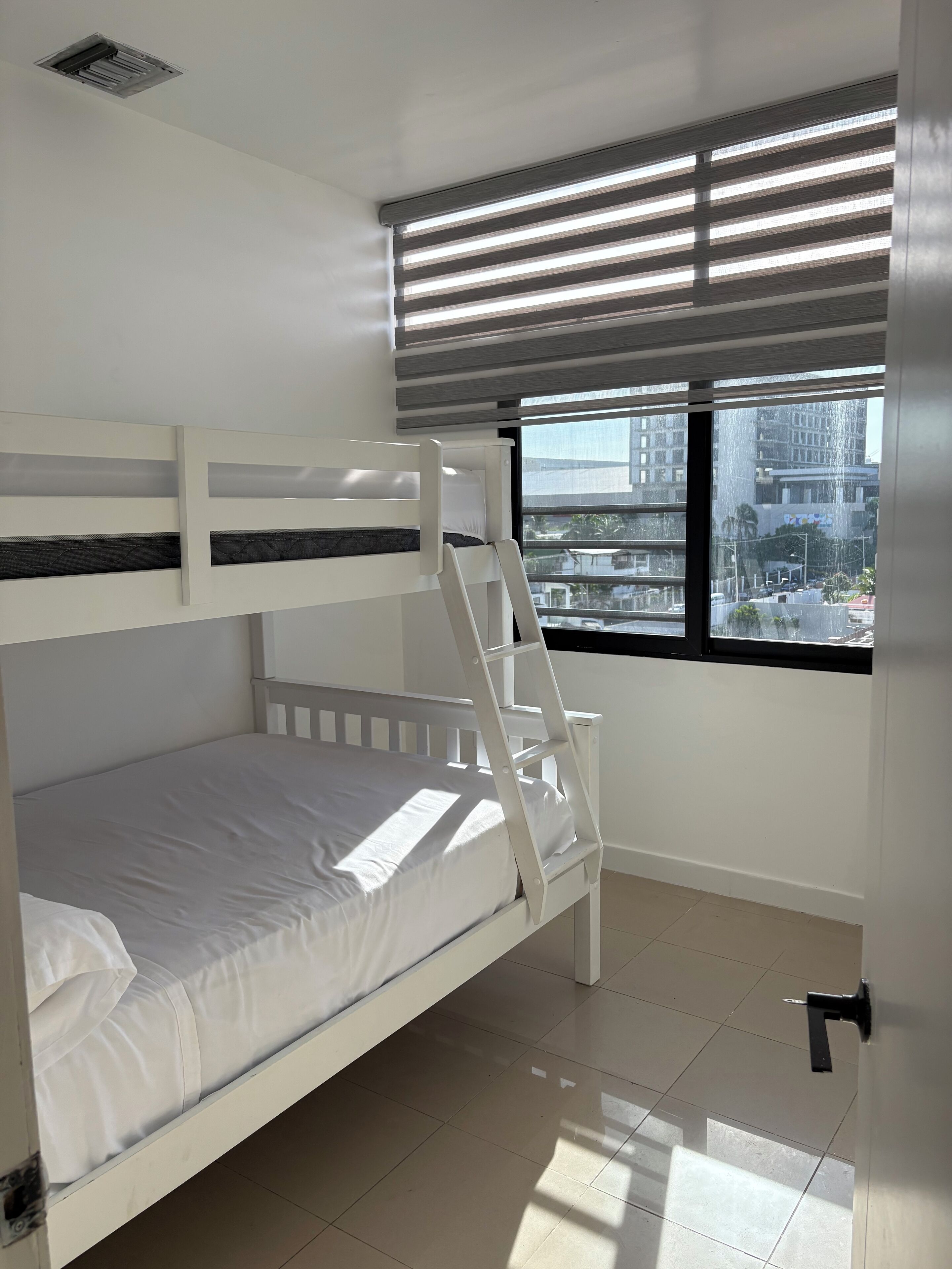 Dormitorio con literas, ventana con persianas, puerta y rejilla de ventilación en el techo.