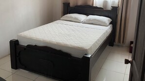 A single bed with a dark frame, a white mattress, and two pillows. There is a ceiling fan, a window with curtains, and a door with a handle.