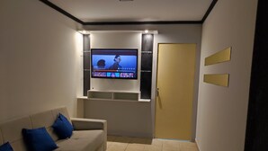 A living room with a flat-screen TV mounted on the wall, a beige sofa with blue cushions, a door, and a tiled floor. - Apartamento Prime Central (Curitiba)