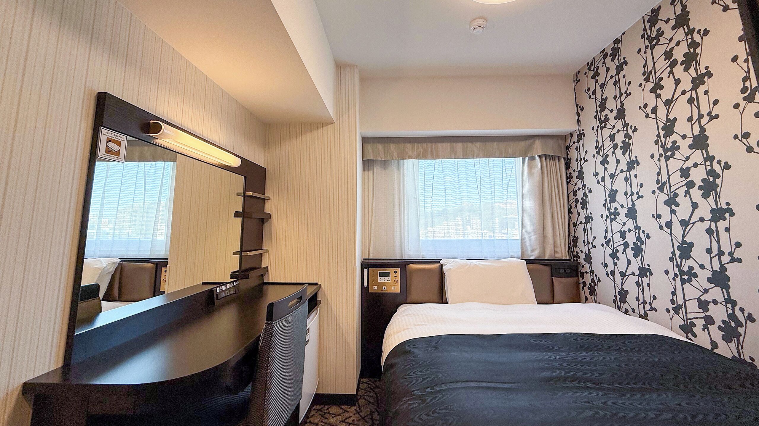 A hotel room with a bed, a desk, a mirror, a window with curtains, and a wall with a floral pattern.