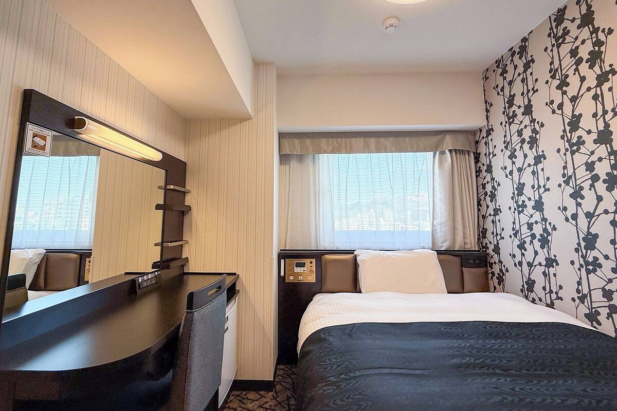 Ein Hotelzimmer mit Bett, Schreibtisch, Spiegel, einem Fenster mit Vorhängen und einer Wand mit Blumenmuster.