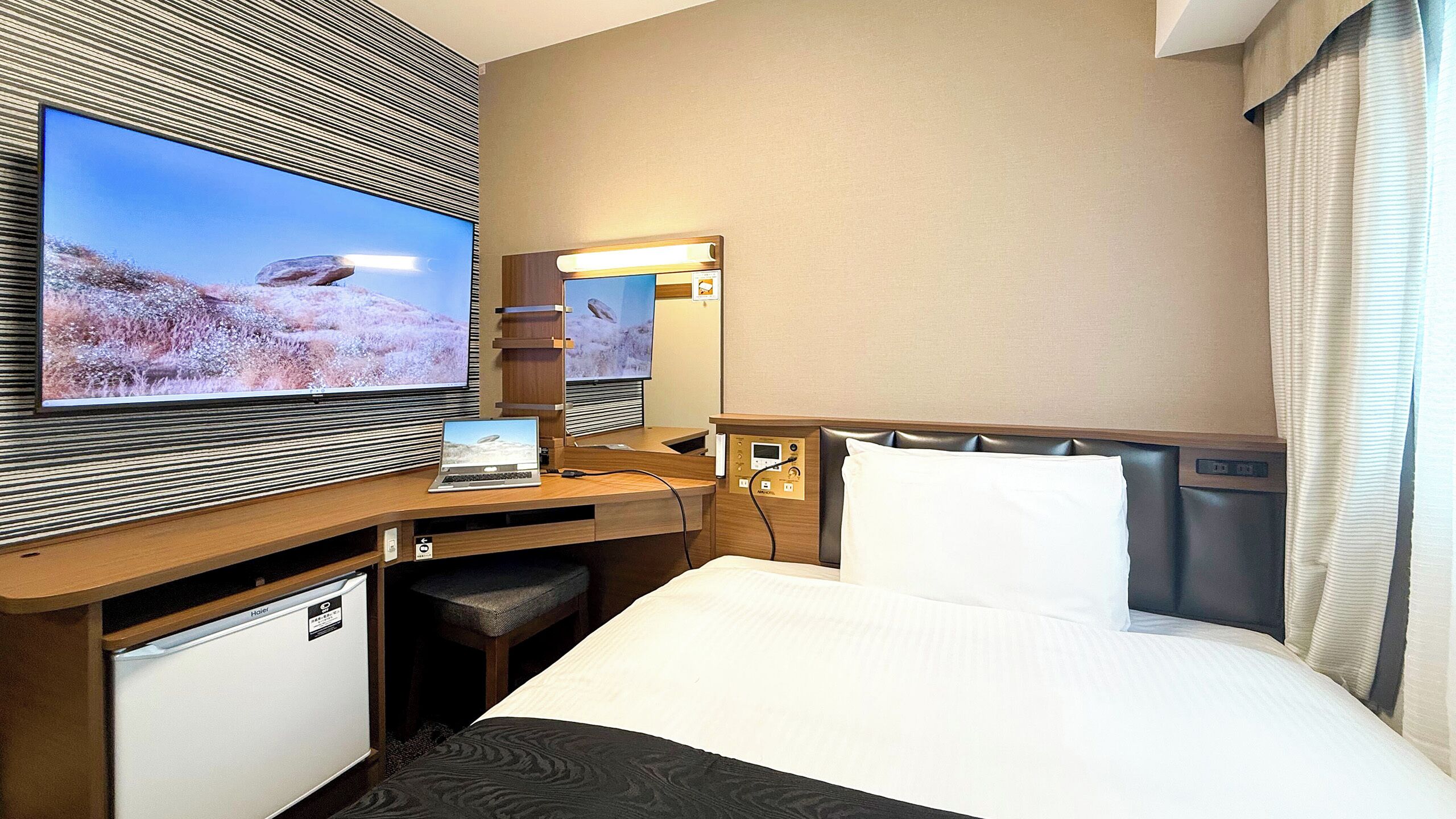 A hotel room with a bed, a desk with a laptop, a television, and a minibar.