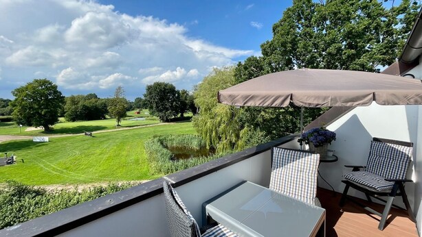 A balcony with a table and chairs, overlooking a golf course and a pond. - 3003 "maison Fleur-de-lis" - Wohnung 3003 am Schoor in Grömitz Inkl. Schwimmbadnutzung (Grömitz)