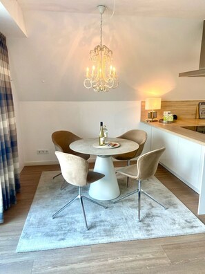 A modern dining area with a round table, four chairs, a chandelier, and a kitchen counter with a lamp. - 3003 "maison Fleur-de-lis" - Wohnung 3003 am Schoor in Grömitz Inkl. Schwimmbadnutzung (Grömitz)