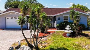 A single-story house with a garage, surrounded by palm trees and a small garden with a fountain.