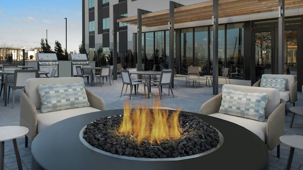 Un área de estar exterior moderna con una chimenea central, rodeada de mobiliario de mimbre y un edificio de vidrio al fondo.