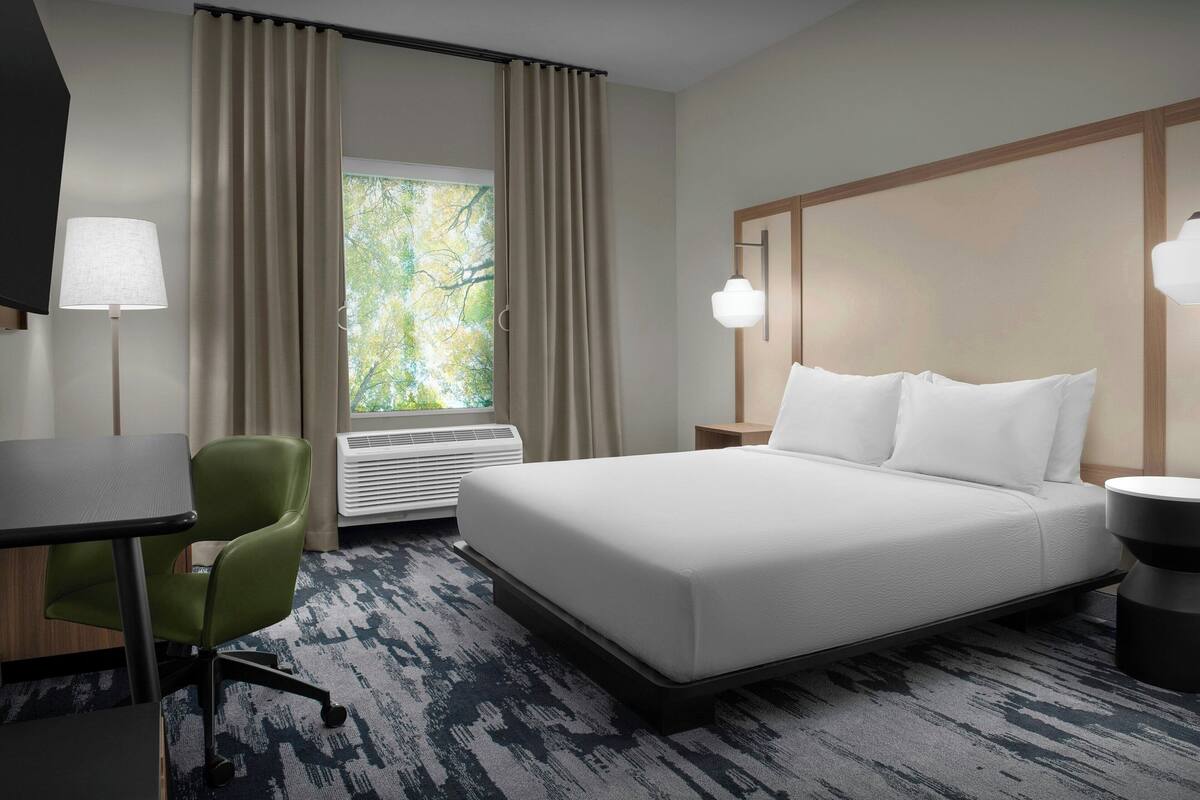 A hotel room with a bed, a desk, a chair, a window with curtains, and a wall-mounted lamp.