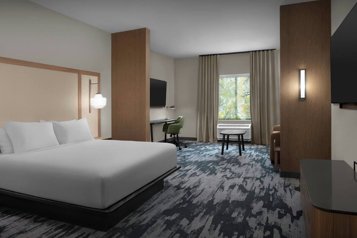 A hotel room with a large bed, a desk with a chair, a TV mounted on the wall, and a window with curtains.