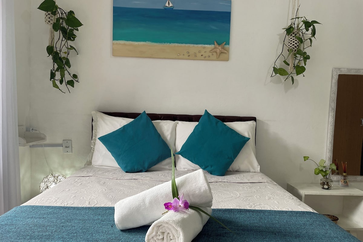 Un lit bien fait, aux couleurs turquoise et blanche, avec un tableau représentant une scène de plage au-dessus, et une petite plante sur la table de chevet.