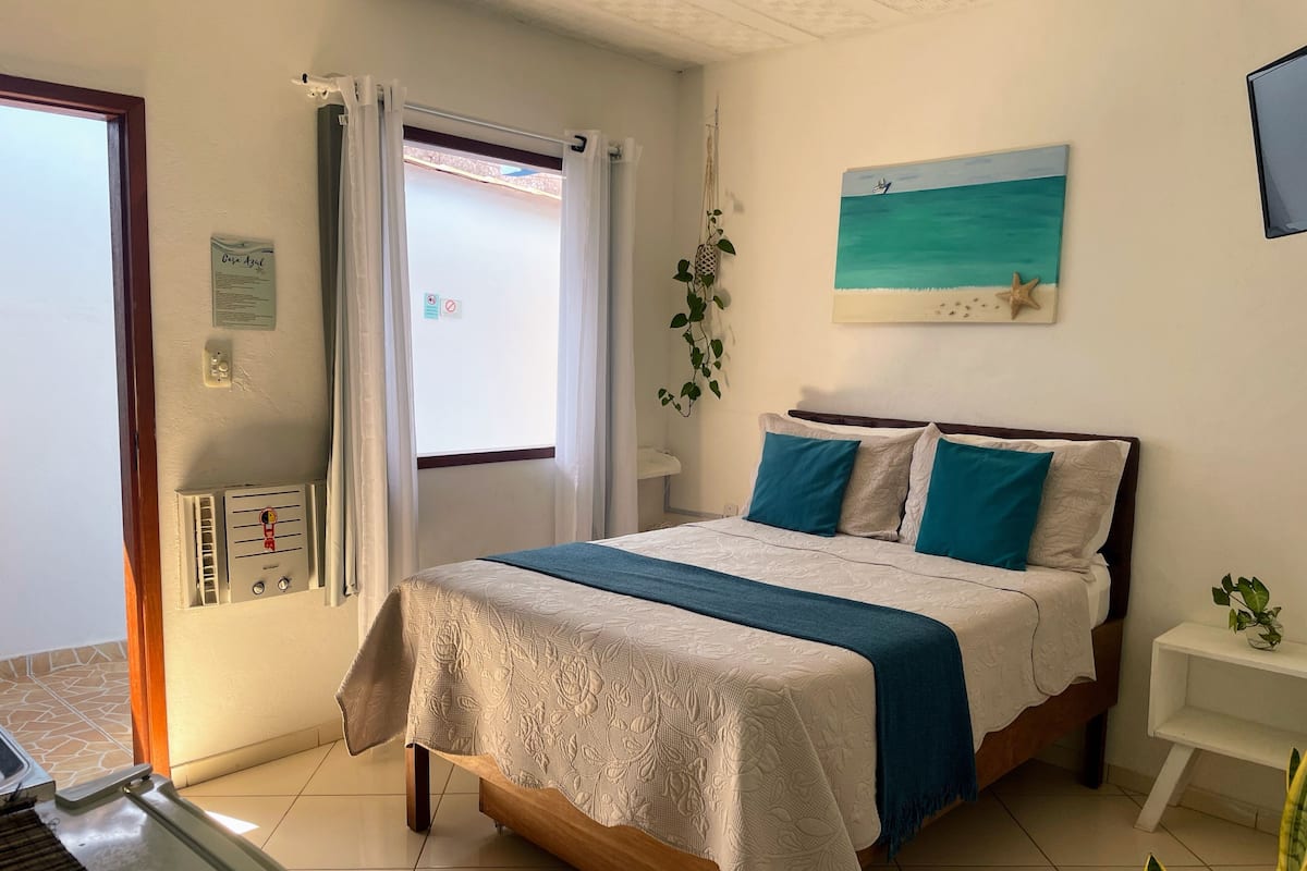 Une chambre à coucher bien rangée, avec un lit, une table de chevet, un tableau accroché au mur et une fenêtre avec des rideaux.
