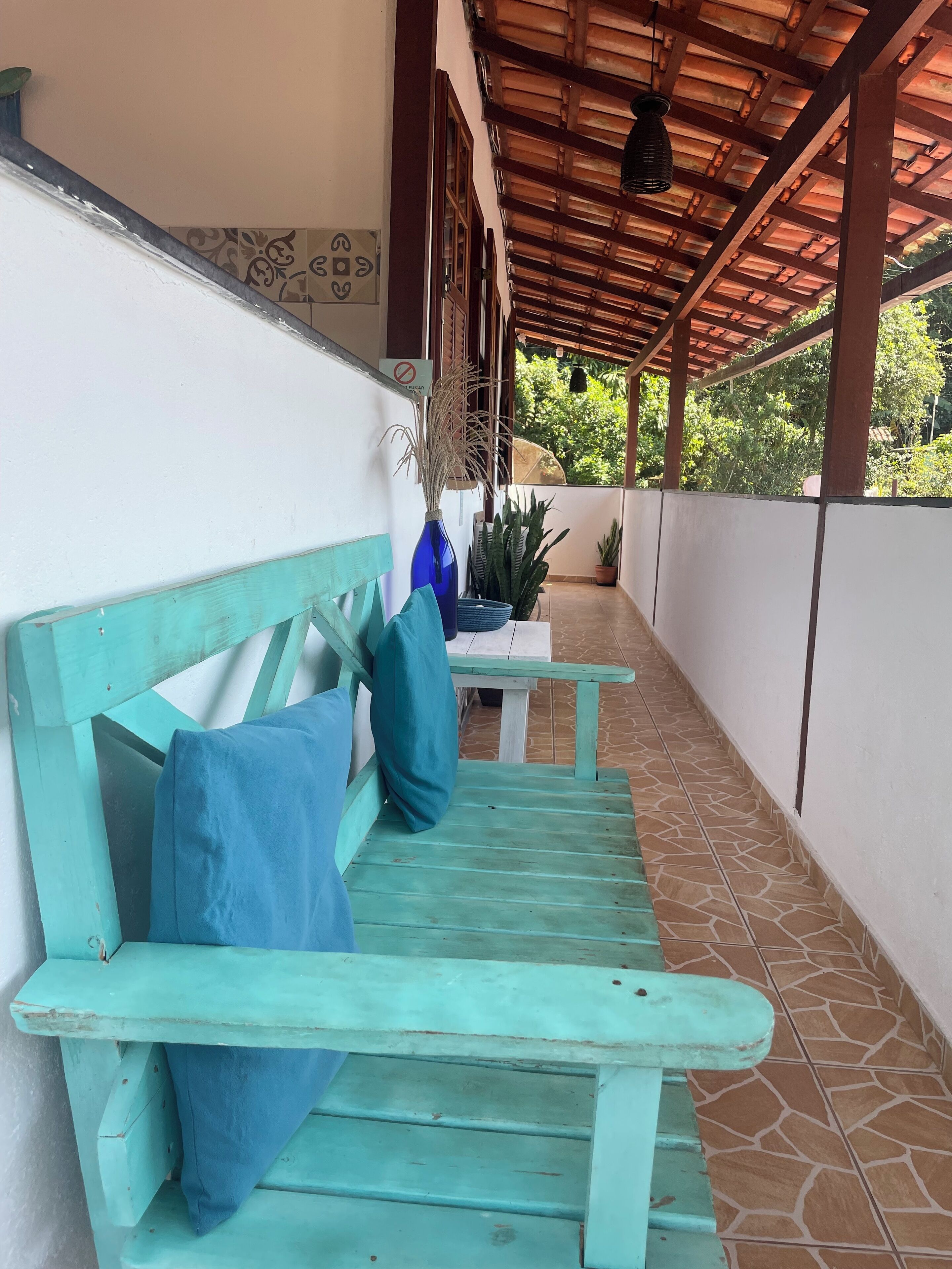 Un espace extérieur aménagé, abrité et doté d’un banc en bois et d’une table, entouré de plantes en pot et recouvert d’un sol carrelé.