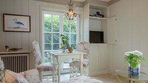 A cozy room with a white floral sofa, a small table with a vase of flowers, a glass coffee table, and a framed picture of a bird on the wall. - Liiger Wal - Typically Frisian: Apartment "Liiger Wal" in Morsum (Morsum)