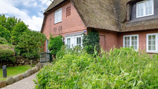 A traditional red brick house with a thatched roof, a number '1968' on the wall, and a well-maintained garden with a stone pathway. - Liiger Wal - Typically Frisian: Apartment "Liiger Wal" in Morsum (Morsum)
