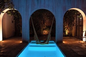 A hammock under a blue-lit archway with a brick wall and plants in the background.