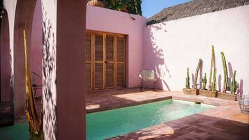 Zona de piscina con pared rosa, contraventanas de madera, una silla y cactus.