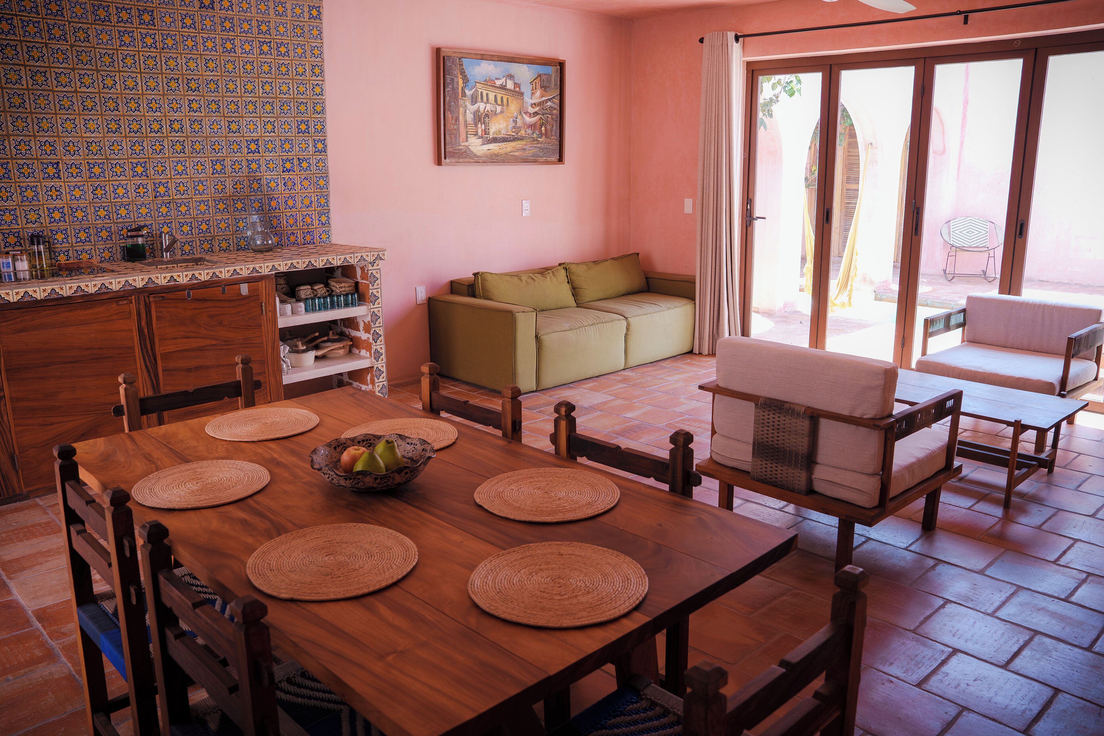 A dining area with a wooden table, chairs, and a bowl of fruit. There is a sofa and a chair in the background.