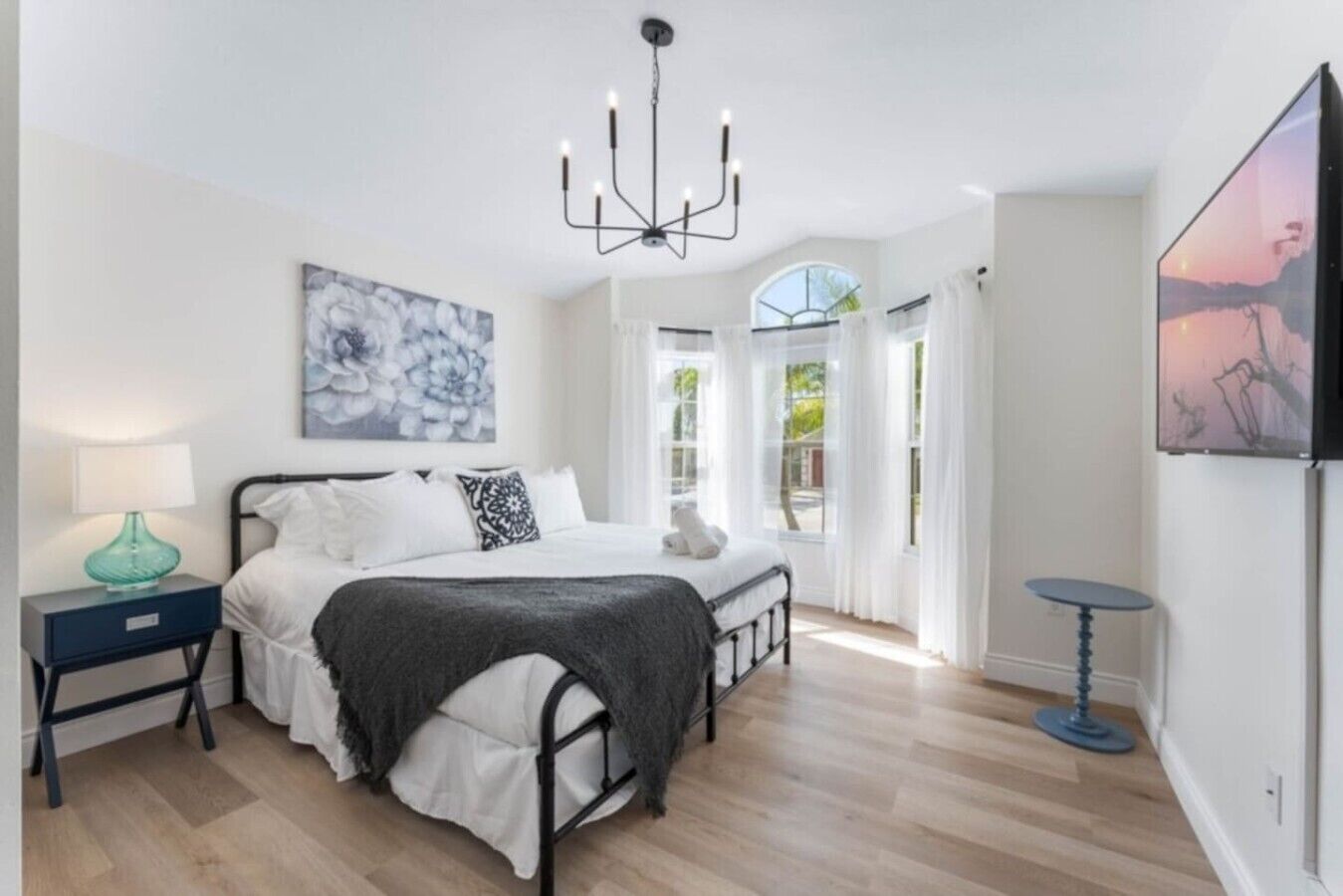A bedroom with a bed, bedside tables, a chandelier, artwork, and a view through a window.