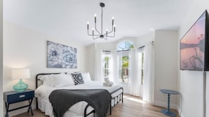 A bedroom with a bed, bedside tables, a chandelier, artwork, and a view through a window.