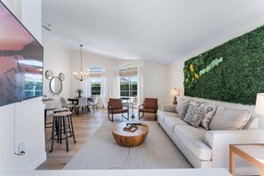 A modern living room with a sofa, coffee table, and chairs. There is a dining area with a table and chairs. A green wall with sunflowers decorates one side.