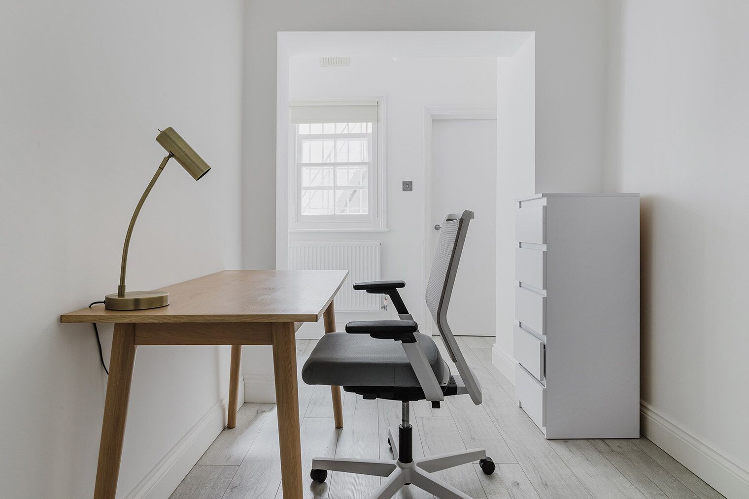 Un espacio de oficina minimalista con un escritorio de madera, una silla moderna, una lámpara y un archivador blanco.