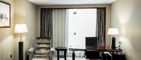 A hotel room with a bed, a desk, two chairs, a lamp, and a large window with curtains.